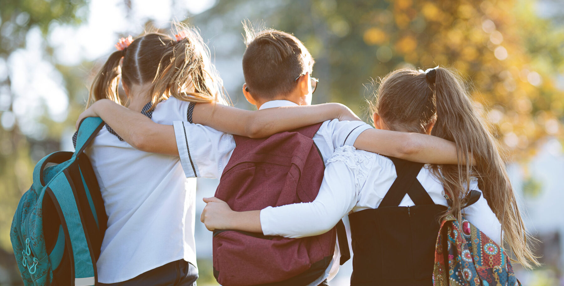 Three children wearing backpacks walk closely together with arms around each other outdoors in daylight.