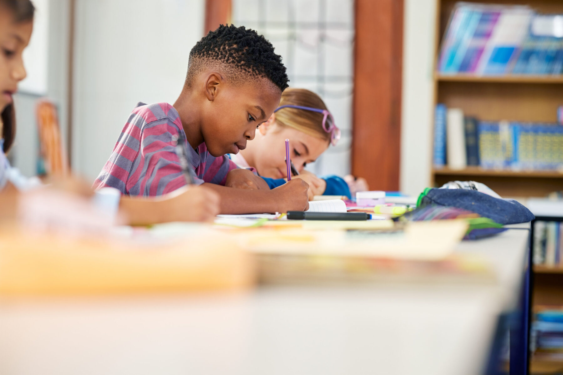 Several children focus on writing at a desk inside a classroom, with school supplies spread out around them.