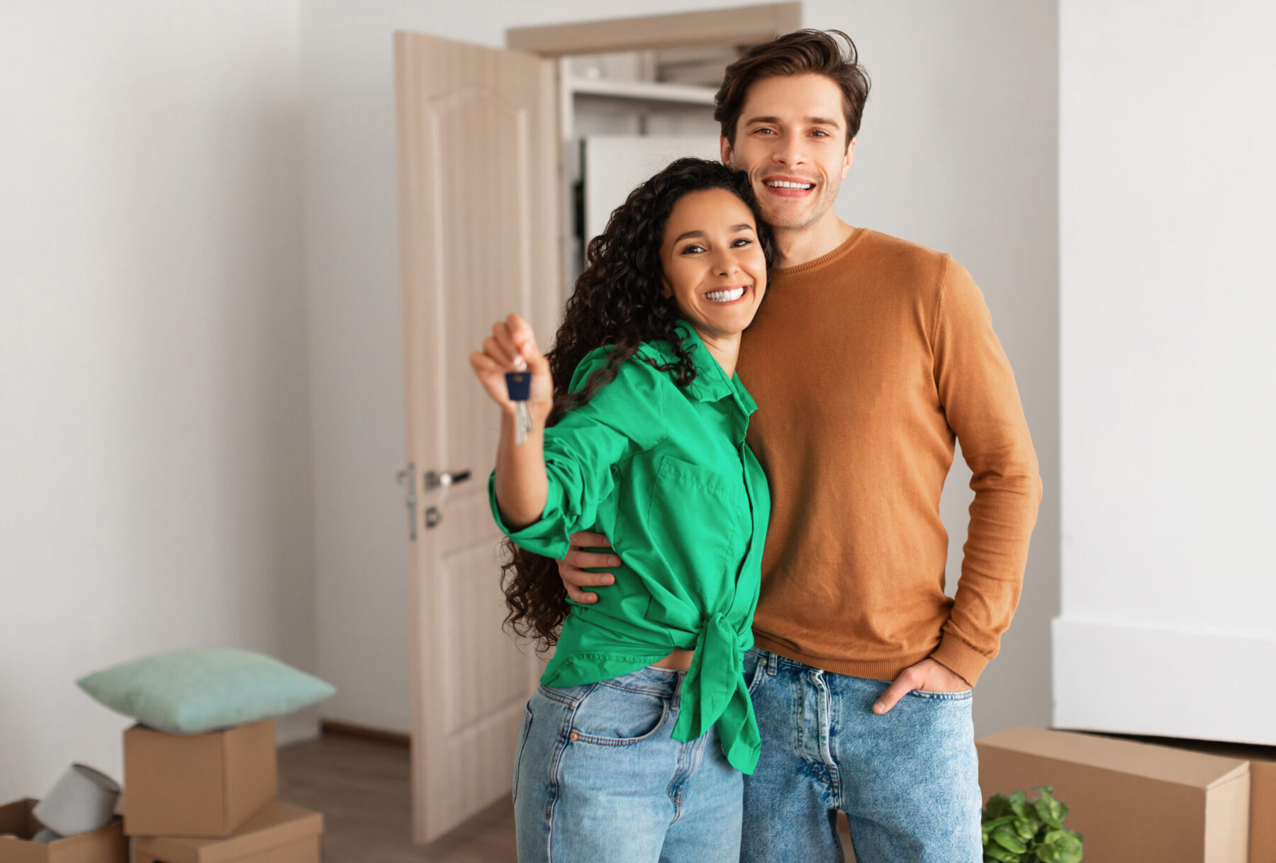 A smiling couple stands in a bright room holding a key, with cardboard boxes in the background.