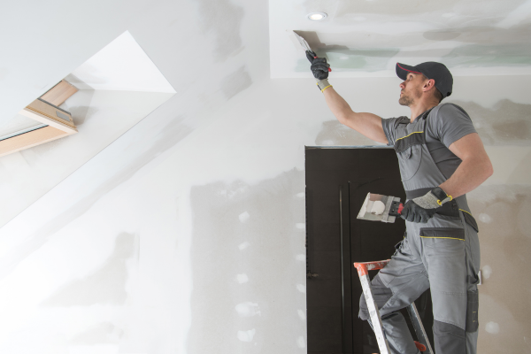 A contractor is standing on a ladder while applying joint compound or plaster to a ceiling during a home renovation. The room appears to be under construction, with unfinished drywall, patch marks on the walls, and a skylight bringing natural light into the space. The image shows an active remodeling or repair project, likely part of interior finishing work before painting.
