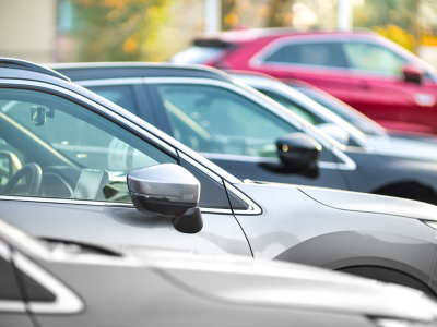 A close-up view of several parked cars lined up in a row, with side mirrors and windows visible in the foreground. A red vehicle stands out among the silver and white cars, and the background is blurred with warm outdoor lighting.