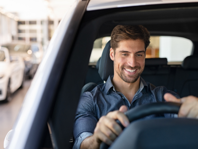 A man wearing a blue button-up shirt sits in the driver's seat of a car, smiling while holding the steering wheel. The interior of the vehicle and a blurred outdoor scene through the windshield are visible in the background.