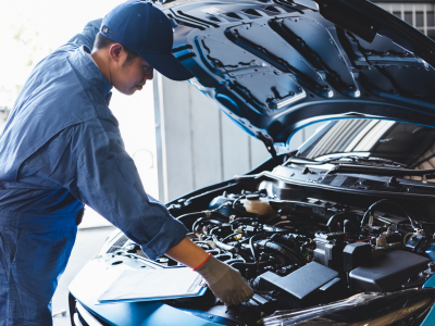 A mechanic wearing a dark blue uniform, cap, and orange gloves leans over the open hood of a car, examining the engine. The workshop setting is visible in the background.