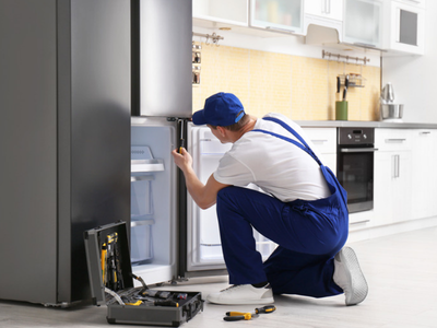A technician in a blue cap and overalls kneeling beside an open toolbox, using a screwdriver inside a refrigerator.