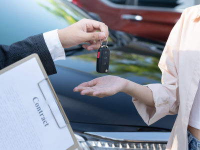 A close-up of a hand in a dark suit sleeve passing a set of car keys with a black remote fob to an open palm. A clipboard labeled "Contract" is visible in the foreground, with a vehicle hood behind.