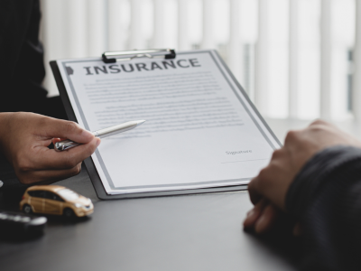 A close-up of a clipboard holding a document titled "Insurance" with a signature line, held by a person pointing at it with a pen. A yellow toy car and a set of keys sit on the desk, with another person's hand resting nearby.