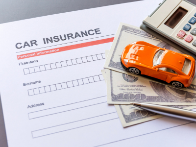 A close-up of a car insurance application form with fields for personal information. An orange toy car, several U.S. dollar bills, a pen, and a calculator are placed on top of the document.